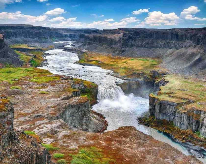 Hafragilsfoss Waterfall plunges dramatically into a rugged canyon, surrounded by rocky cliffs and vibrant patches of green, under a bright blue sky with scattered clouds.