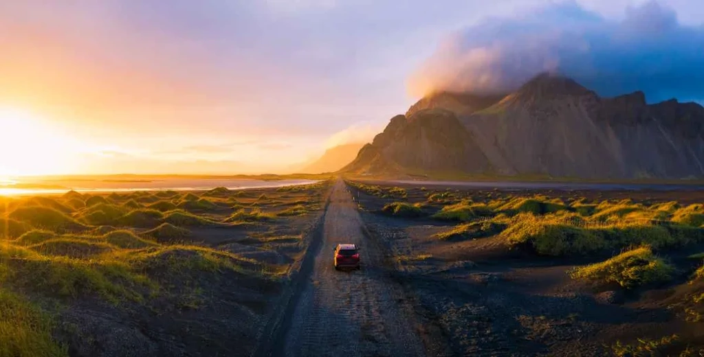 Red car driving along a remote gravel road in Iceland at sunset, with green mossy terrain and dramatic mountains rising beside the coast.