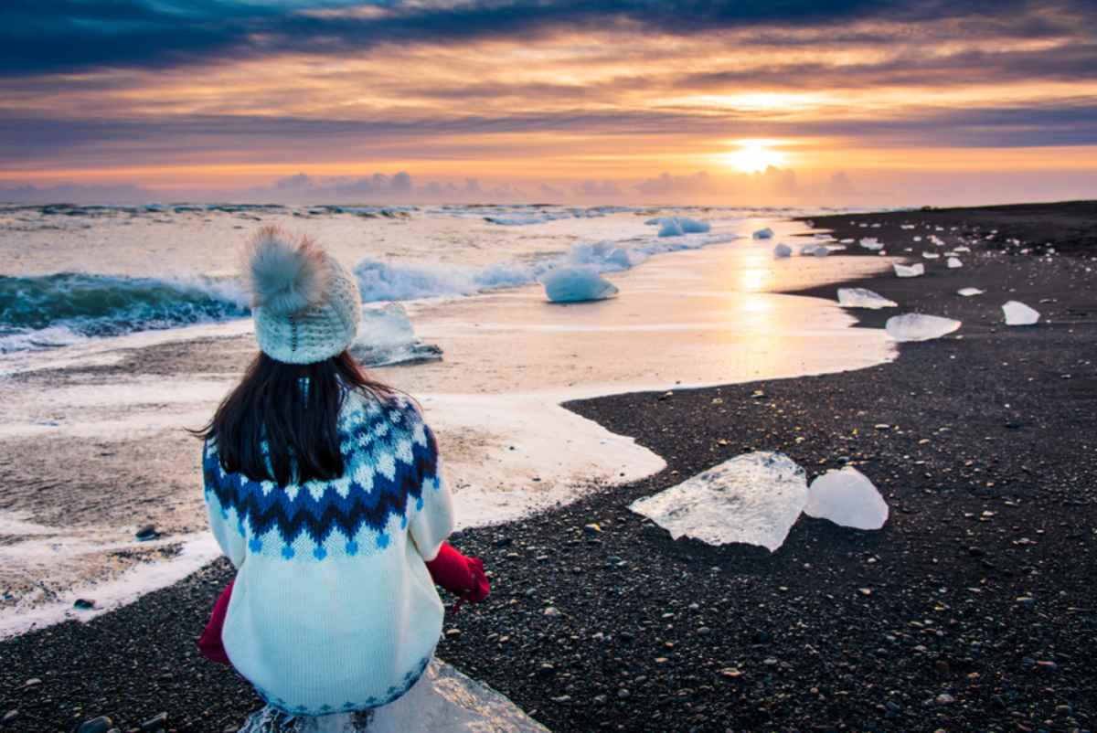 A sheltered woman sits on the shore of Iceland's Diamond Beach as she watches the waves crash over the icy rocks and the sunset.