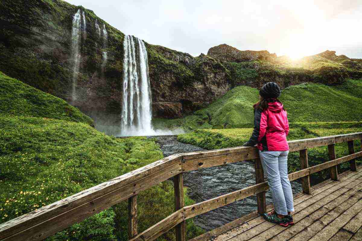A brunette woman with long hair watches the water fall from the Seljalandsfoss waterfall in Iceland respecting the safety perimeter marked by its viewpoint.