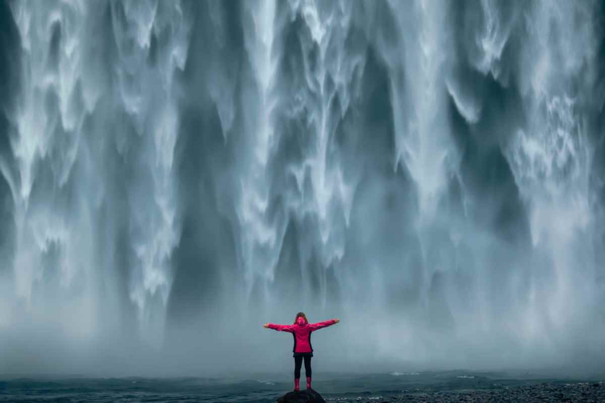 A woman in a fuchsia raincoat stands in the center of the image while the spectacular waterfall of Skogafoss waterfall in Iceland is in the background.