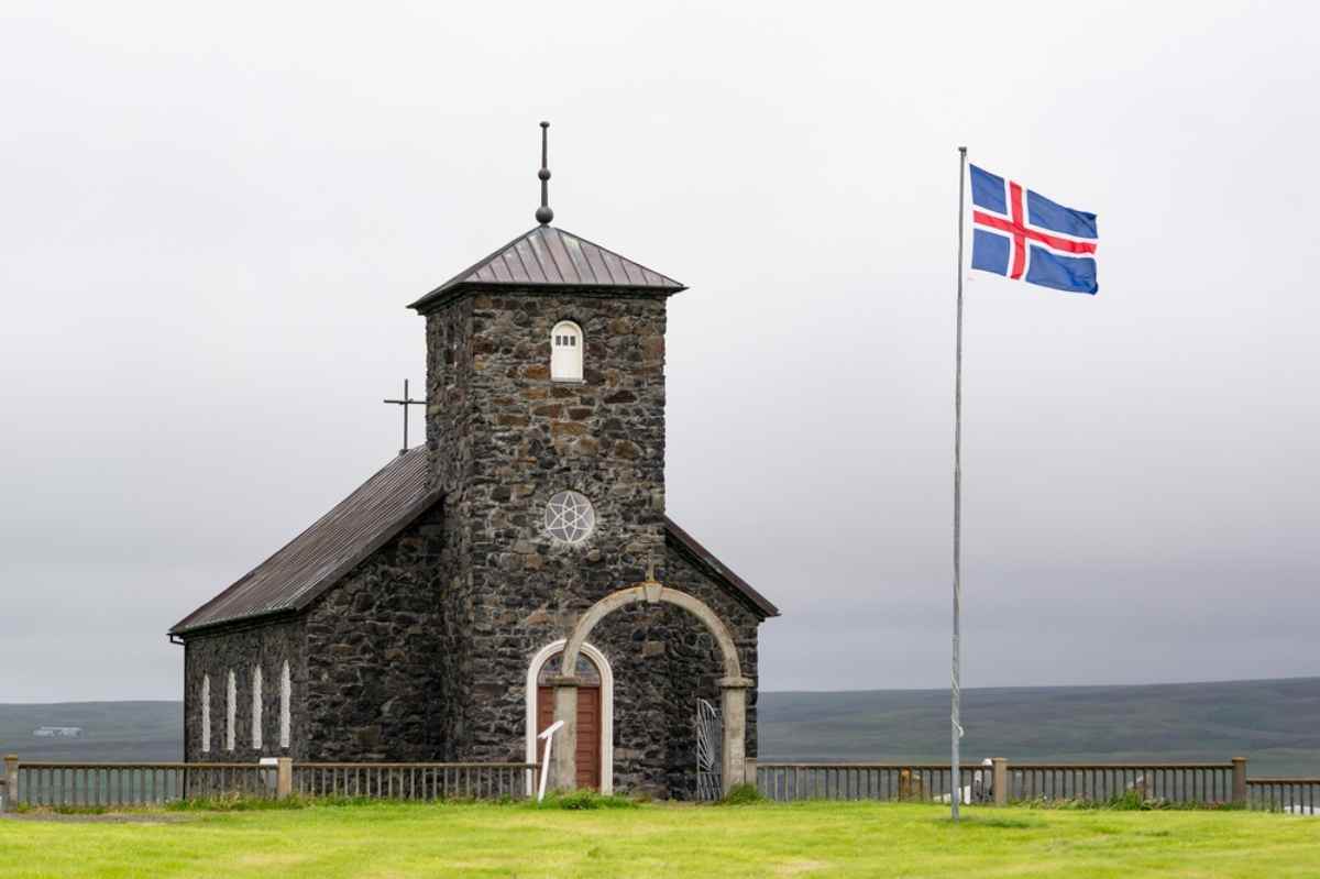 General view of Thingeyrakirkja church in Iceland with a small tower with Christian symbols and a flag of the country.