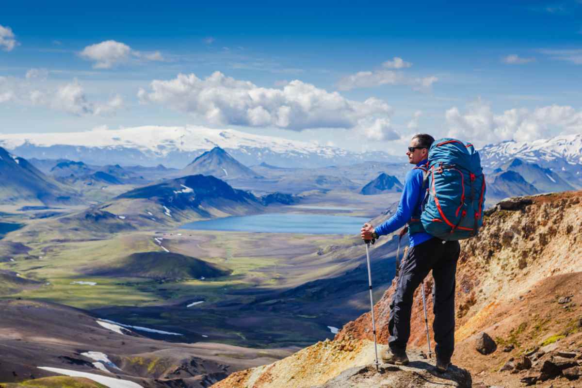 A hiker with a backpack on his back and a walking stick stands motionless at the top of a canyon overlooking a rocky, mountainous Icelandic landscape.