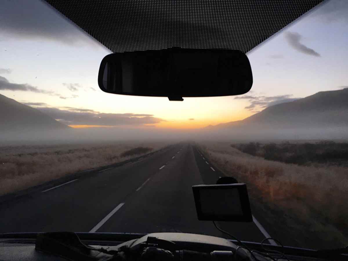 A road at sunset is observed from inside a car with a camera hanging from the window and a rearview mirror in the center of the car.