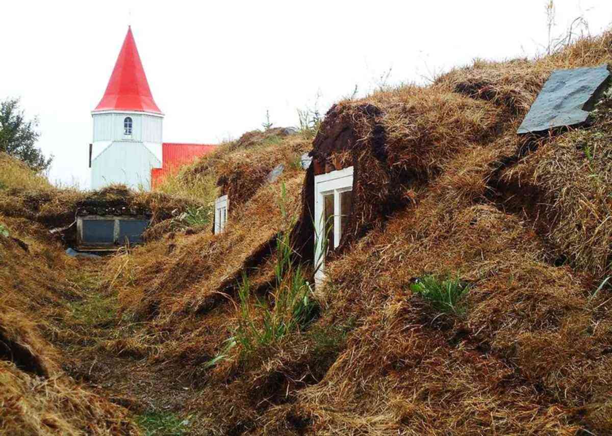 Casas de c&eacute;sped tradicionales islandesas parcialmente enterradas en el suelo con techos cubiertos de hierba, situadas cerca de una iglesia blanca con techo rojo, creando una escena r&uacute;stica e hist&oacute;rica.