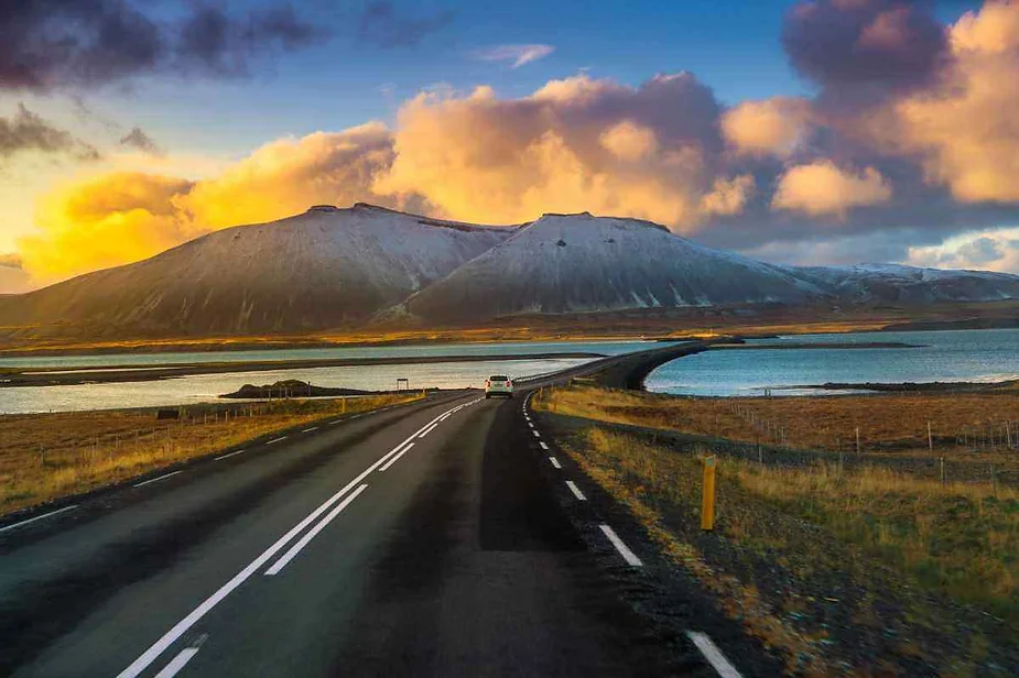 Un recorrido esc&eacute;nico por una carretera islandesa que conduce hacia monta&ntilde;as nevadas, con un vibrante cielo al atardecer y aguas calmadas a ambos lados de la carretera.