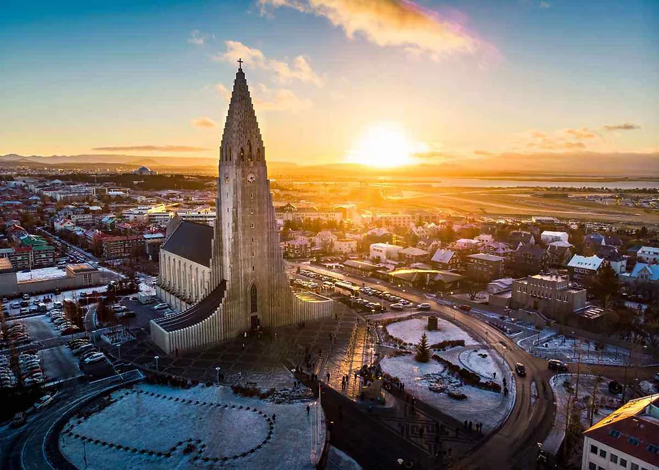 Vista a&eacute;rea de la iglesia Hallgr&iacute;mskirkja en Reikiavik, Islandia, al amanecer, con el sol proyectando una c&aacute;lida luz dorada sobre el paisaje urbano y los tejados cubiertos de nieve.