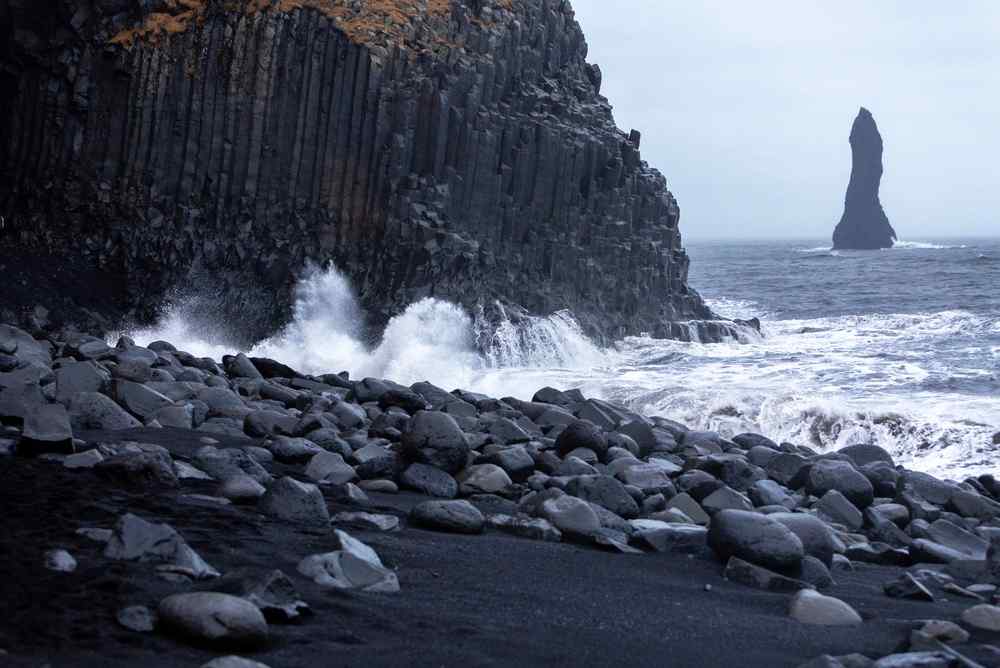 Black sand beach in Iceland Waves crashing against a black sand beach in Iceland near tall basalt cliffs and sea stacks.