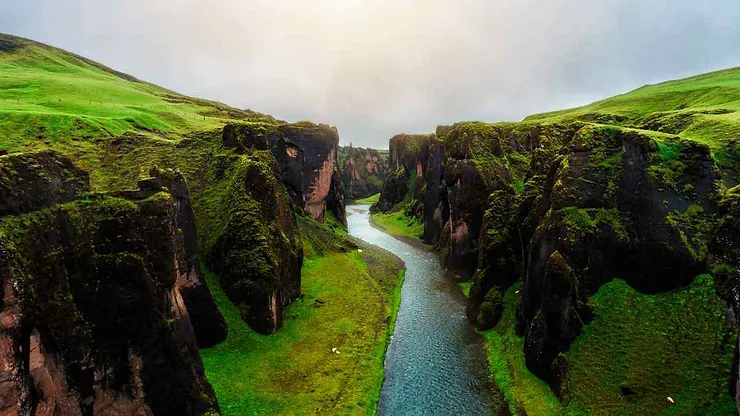 Una vista aerea mozzafiato del canyon di Fjaðrárgljúfur in Islanda, con ripide scogliere ricoperte di muschio e un fiume tortuoso che attraversa il paesaggio verde e rigoglioso.