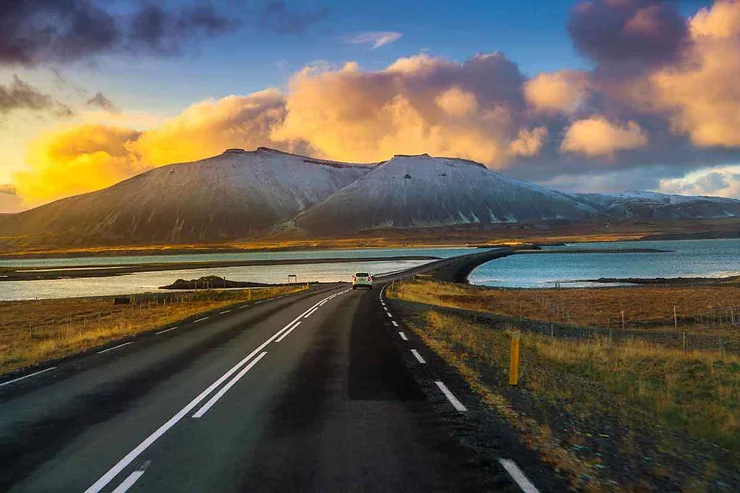 Un viaggio panoramico su una strada islandese che conduce verso montagne innevate, con un cielo al tramonto vibrante e acque calme su entrambi i lati della strada.