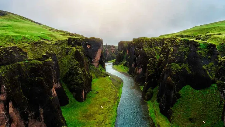 Vue aérienne à couper le souffle du canyon de Fjaðrárgljúfur en Islande, avec des falaises abruptes couvertes de mousse et une rivière sinueuse qui traverse le paysage verdoyant.