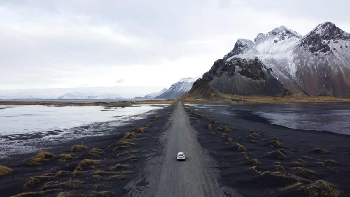 Un coche blanco circula por una carretera de arena negra y vegetaci&oacute;n en Islandia.