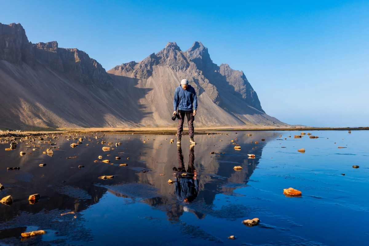 Un homme avec un appareil photo et une veste bleue se refl&egrave;te sur la plage de sable noir devant la montagne Vestrahorn.
