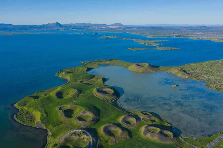 Luftaufnahme des M&yacute;vatn-Sees in Island mit einzigartigen Vulkankratern, &uuml;ppiger gr&uuml;ner Landschaft und dem weiten blauen Wasser des Sees unter einem klaren Himmel.