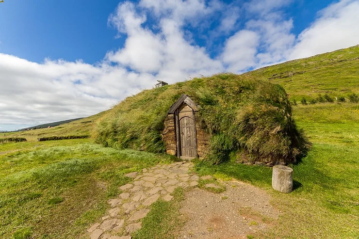 Eiriksstadir A traditional turf house at Eiriksstadir, Iceland, showcasing its rustic wooden door and roof covered with green grass, set against a backdrop of rolling hills and a blue sky with fluffy clouds.