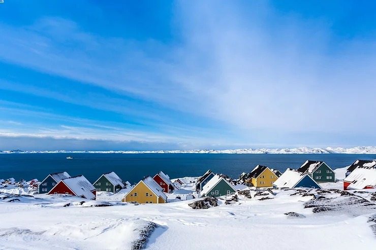 Winter Serenity in Iceland A picturesque view of colorful houses nestled in a snowy landscape by the sea, with a clear blue sky and snow-covered mountains in the distance.