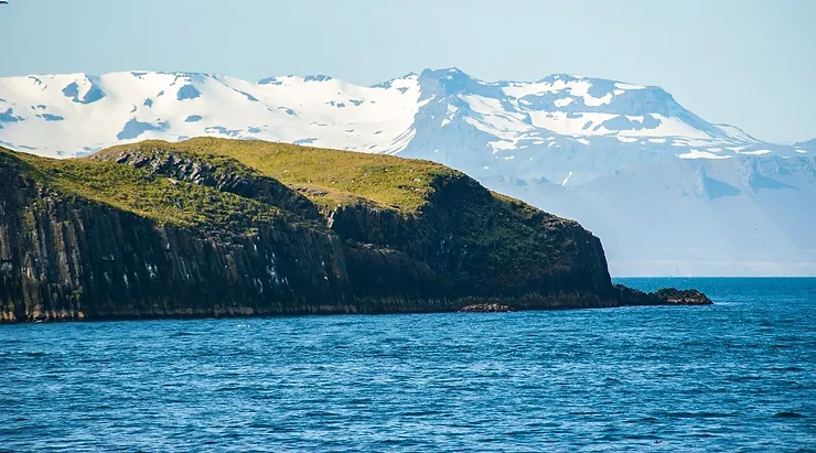 Oxney Island Oxney Island with its rugged coastline and green grass, set against the backdrop of snow-capped mountains under a clear blue sky.