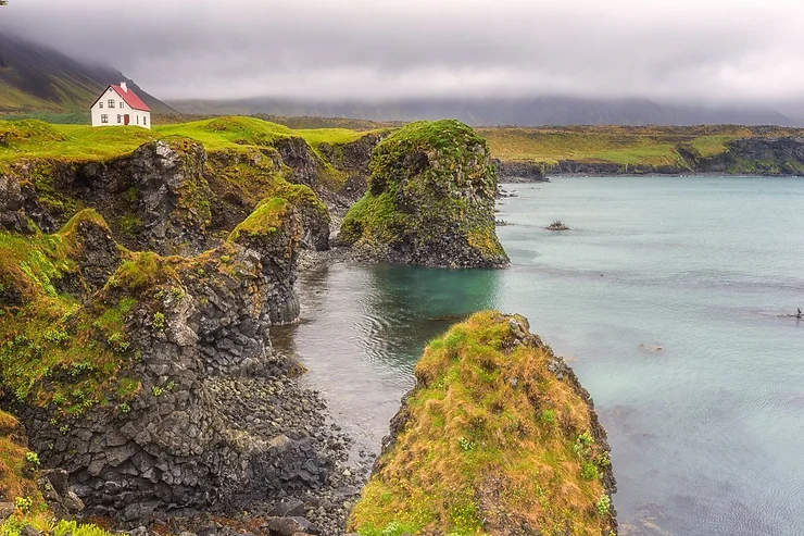 Snæfellsnes Peninsula A charming house perched on the green cliffs of Snæfellsnes Peninsula, overlooking the tranquil turquoise waters, with a backdrop of misty mountains.