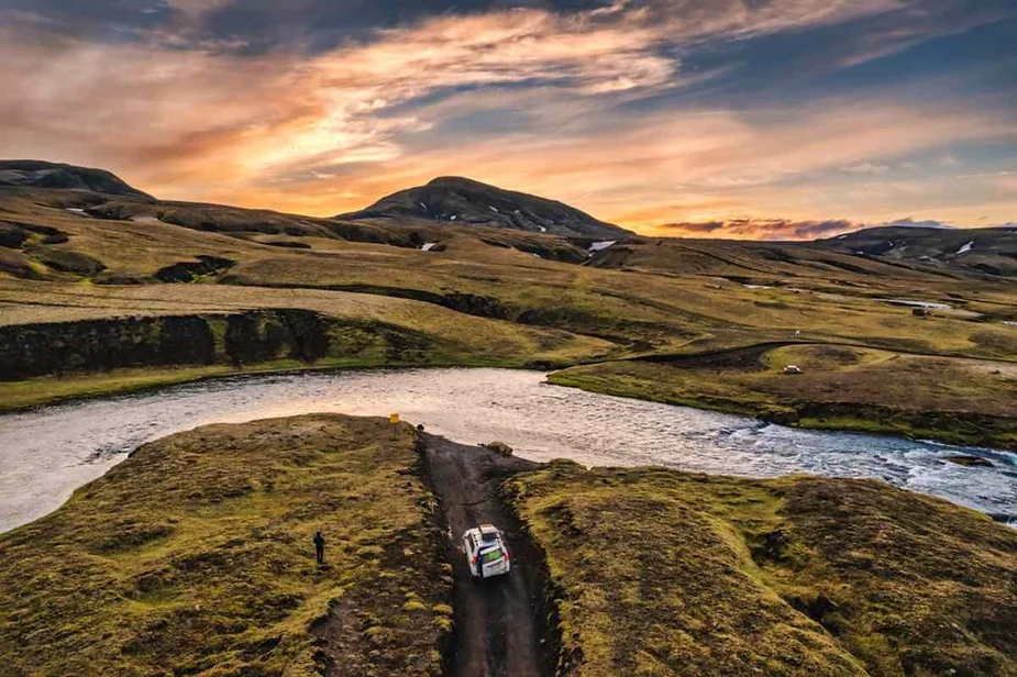 Aventurándote por las Tierras Altas de Islandia al Atardecer: Una Guía para Elegir el Perfecto 4x4 Un vehículo 4x4 recorre un camino de tierra en las remotas Tierras Altas de Islandia al atardecer, cruzando un río entre colinas onduladas y un cielo dramático. La escena enfatiza el espíritu aventurero de los viajes todoterreno por la naturaleza salvaje de Islandia.