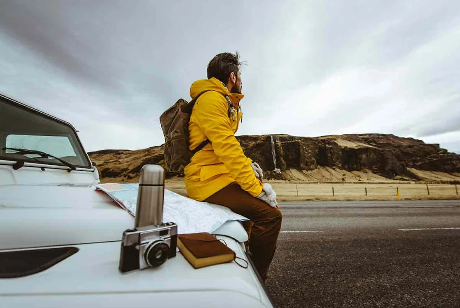 Una viaggiatrice in giacca gialla è seduta sul cofano di un'auto parcheggiata su una strada islandese, con una mappa, una macchina fotografica e una borraccia accanto a lei. Il paesaggio pittoresco, con scogliere e una cascata lontana, sottolinea un'avventura on the road attraverso l'unica bellezza naturale dell'Islanda.