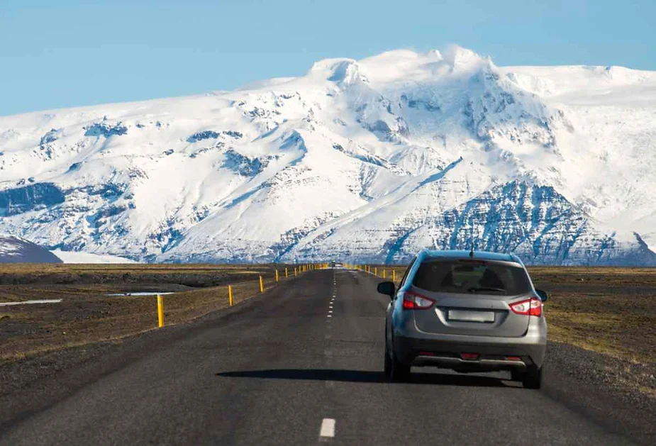Un'auto argentata percorre una strada sgombra verso le montagne innevate dell'Islanda, mostrando il paesaggio drammatico e le condizioni stradali ideali per un road trip in Islanda.