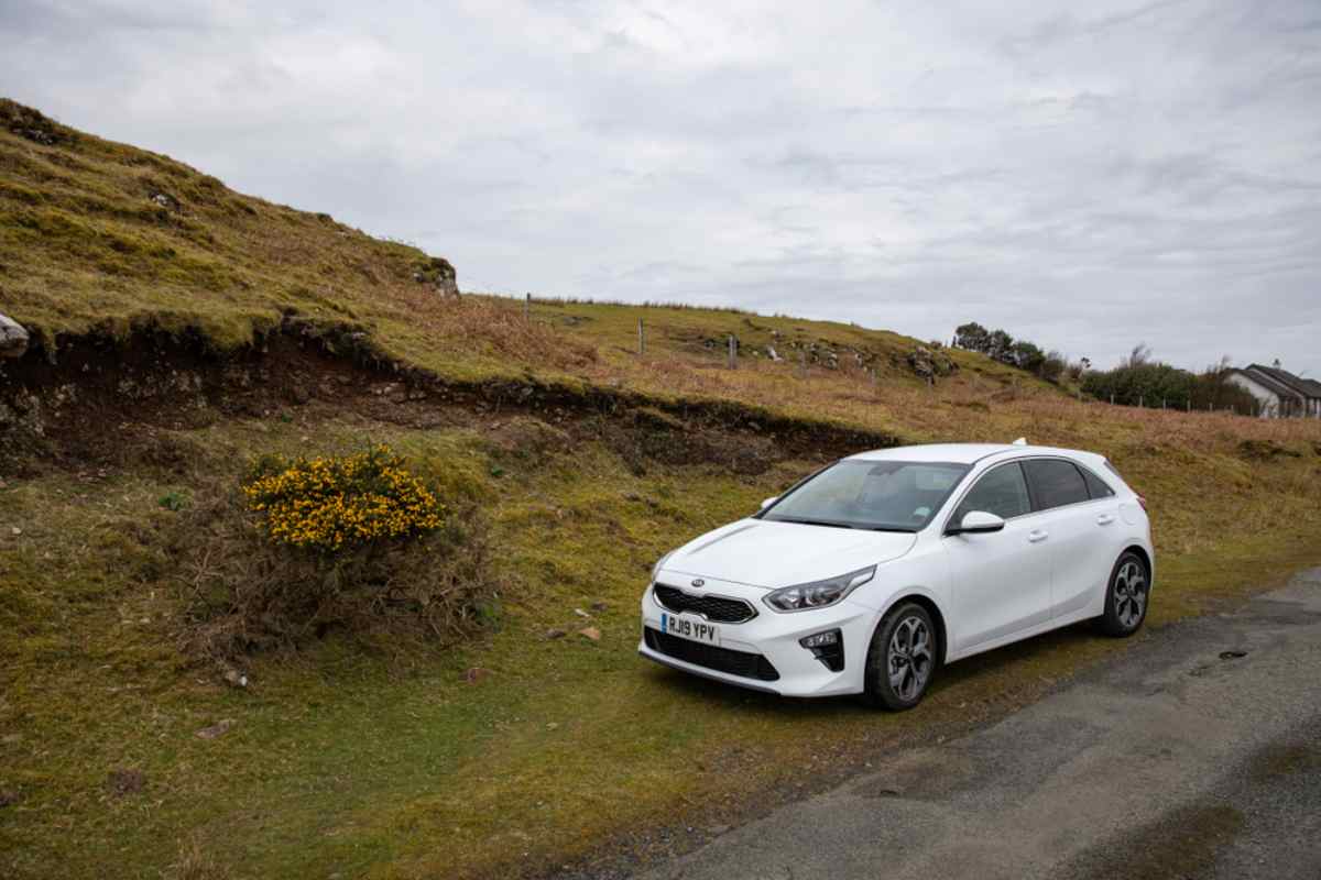 White Kia Ceed parked along a rural road with grassy hills in Iceland.