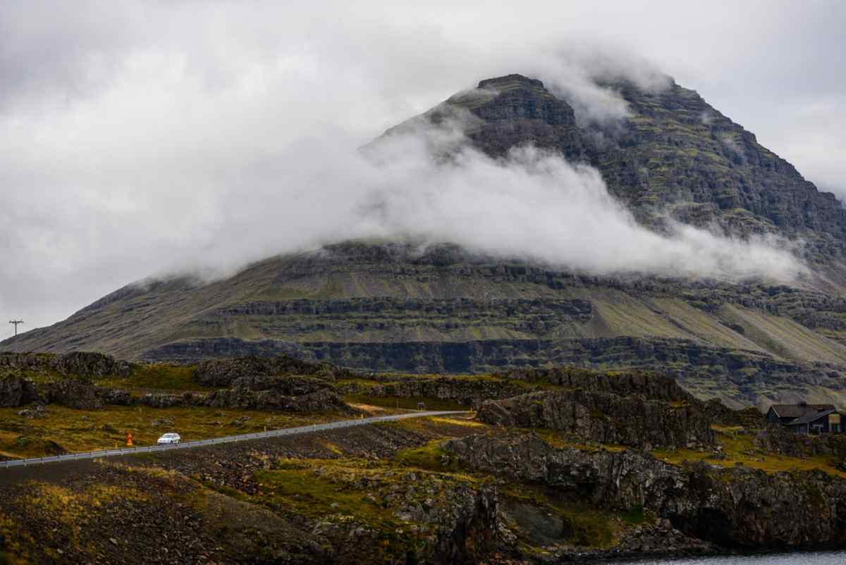 Kleines Auto auf kurviger Straße unter dramatischen Bergen und tiefen Wolken in Islands Ostfjorden.