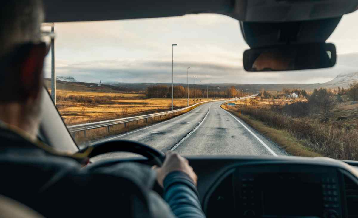 Driver’s view through the windshield on an open road in Iceland, with rolling fields and distant mountains ahead.