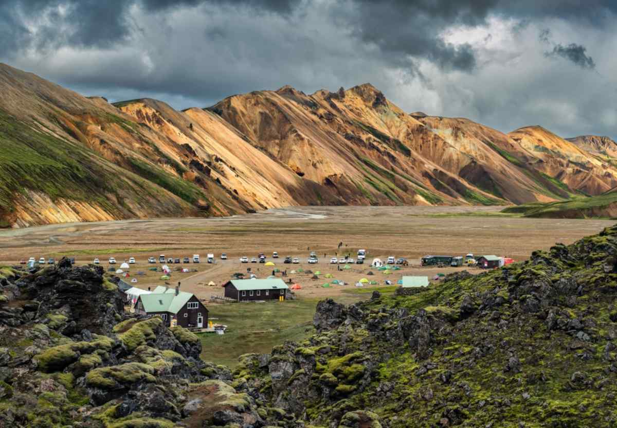 Campsite in Iceland surrounded by colorful volcanic mountains, with tents, cars, and small cabins spread across a wide valley.