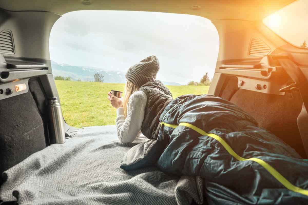 Traveler resting in the back of a car under a sleeping bag, holding a mug and looking out over a grassy landscape at sunrise.