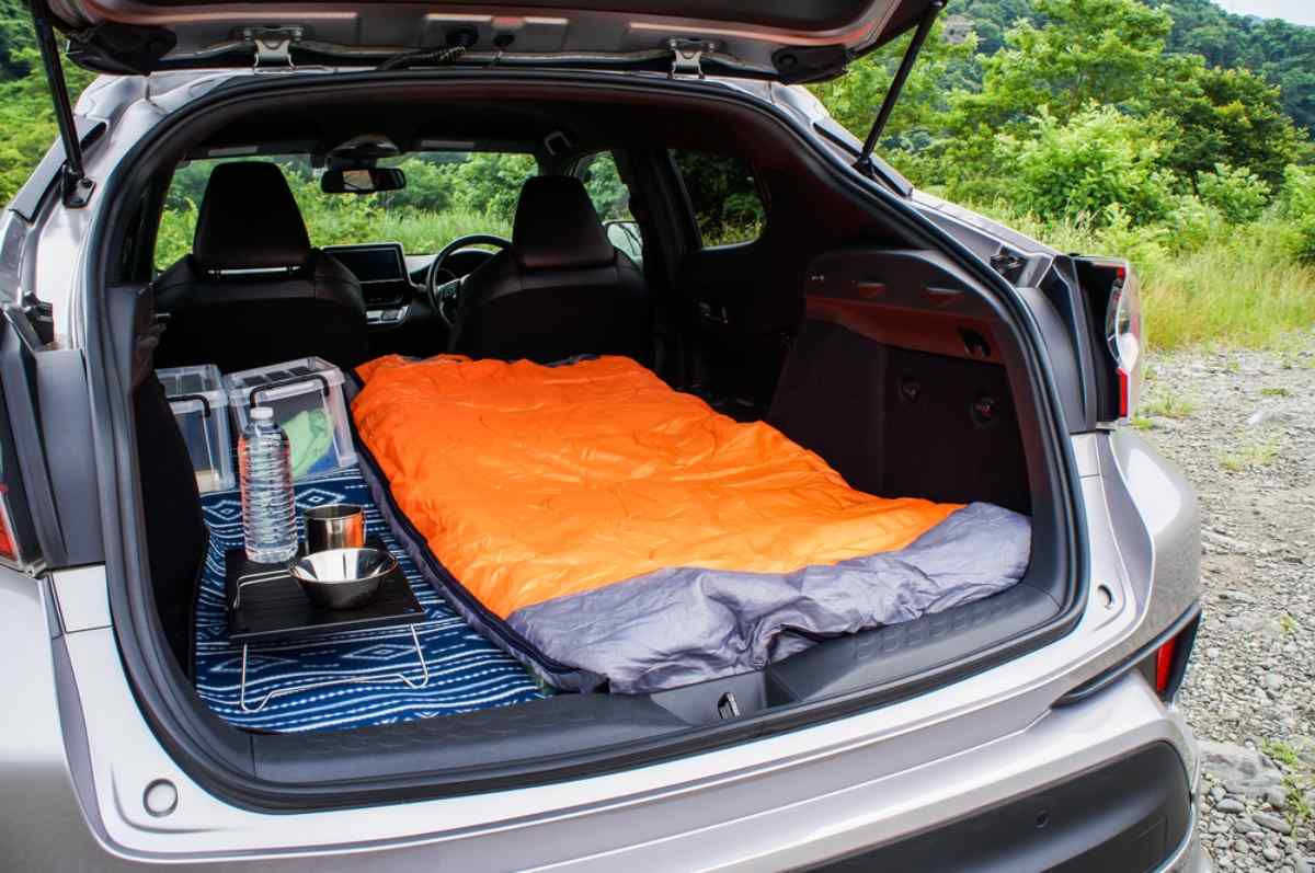 SUV trunk set up for sleeping with an orange sleeping bag, camping gear, water bottle, and small tray on a patterned mat.