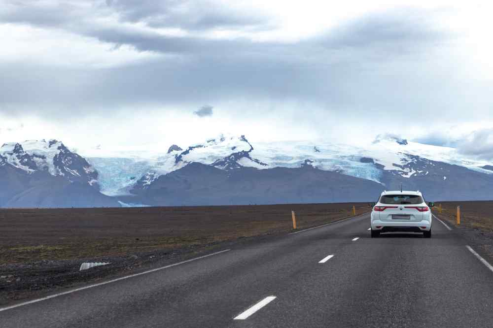 White SUV driving along an Icelandic road with glaciers and mountains in the background.