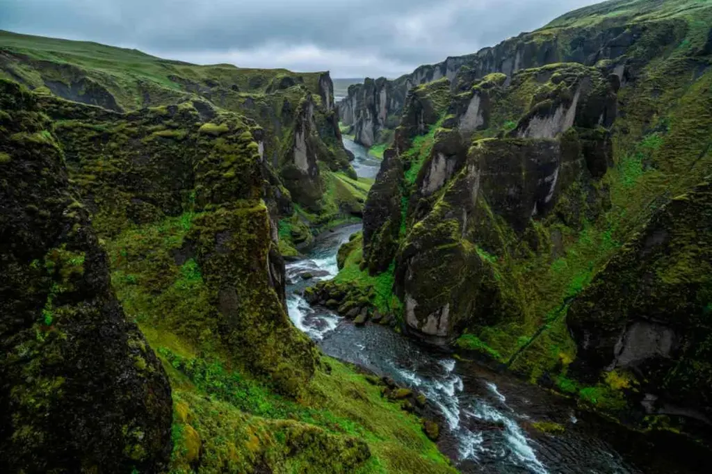A breathtaking view of Fjadrargljufur Canyon in Iceland, showcasing its deep, narrow gorge with steep, moss-covered cliffs. A river flows through the bottom of the canyon, winding its way between the towering rock formations. The lush greenery and the dramatic rock formations create a stunning landscape, with the sky above showing signs of overcast weather. This natural wonder is a testament to Iceland's rugged and untouched beauty, offering an awe-inspiring sight for visitors.