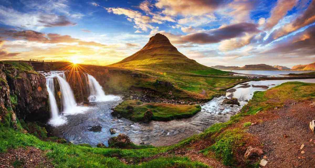Ikonischer Blick auf den Berg Kirkjufell mit dem Wasserfall Kirkjufellsfoss im Vordergrund während eines farbenprächtigen Sonnenuntergangs in Island, wobei die Sonnenstrahlen einen goldenen Schein auf die Landschaft werfen.