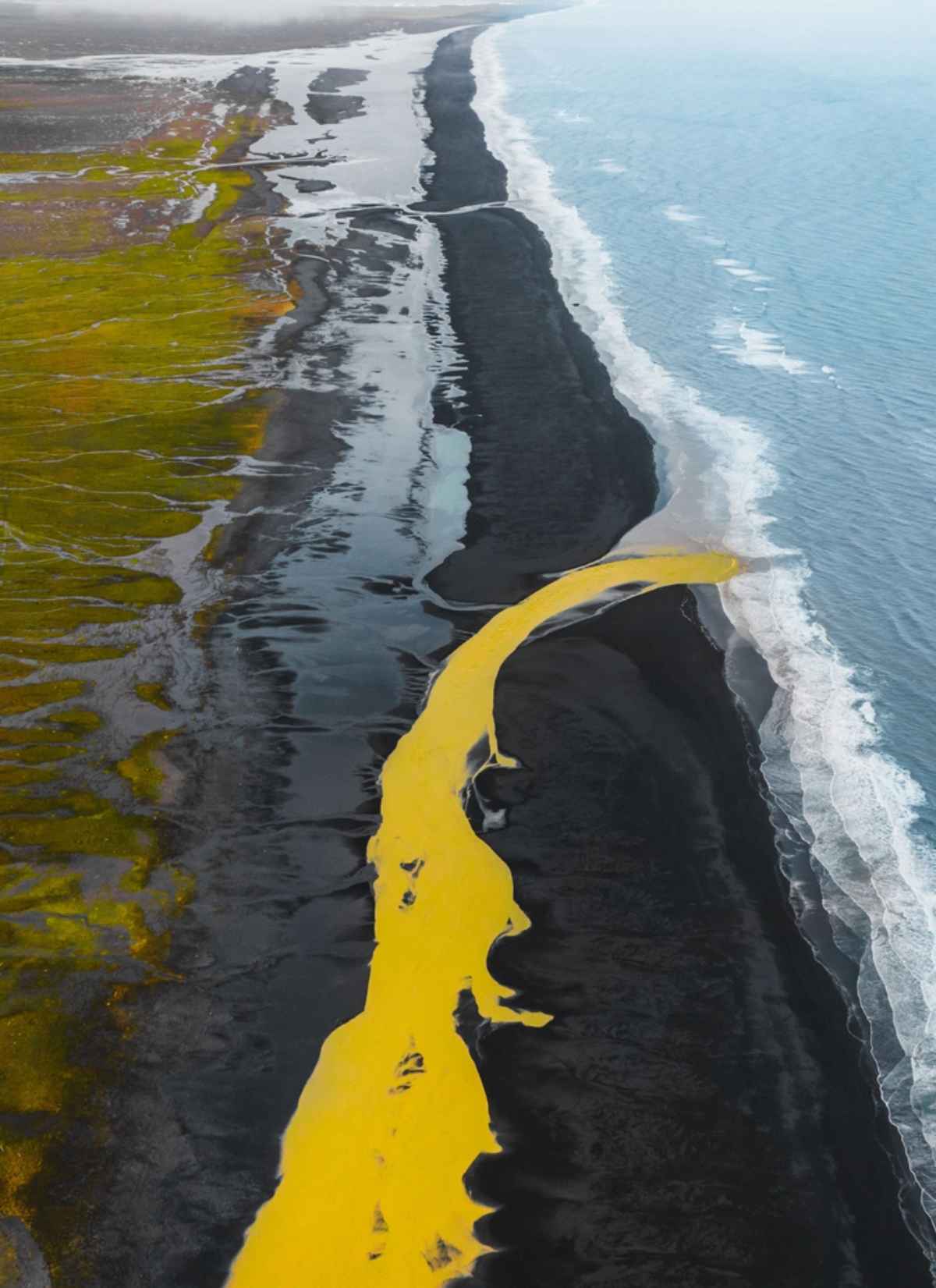 Drohnenaufnahme, kontrastreiche Landschaft, in der der Fluss sich durch den Schwarzsand windet und im Meer in Island mündet