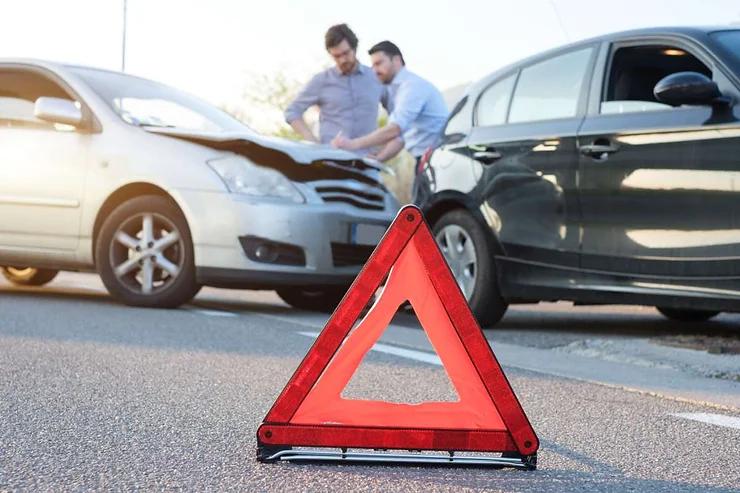 Ein rotes Warndreieck im Vordergrund, während zwei Männer im Hintergrund die Schäden an ihren Autos nach einer Kollision begutachten. Ein Auto ist silber, das andere schwarz, was die Folgen eines Autounfalls zeigt.