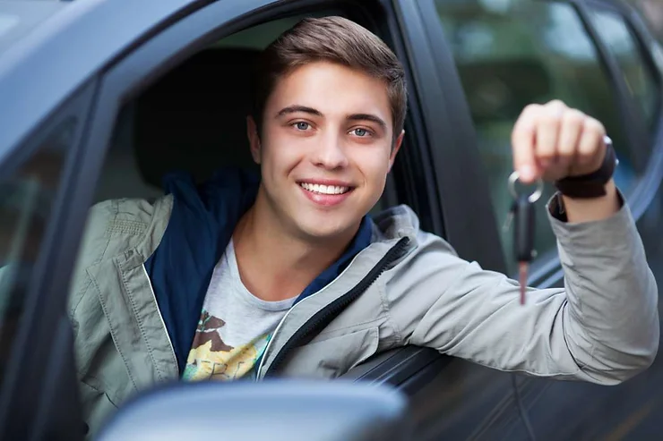 Ein junger Mann sitzt lächelnd auf dem Fahrersitz eines Autos und hält die Autoschlüssel in der Hand.