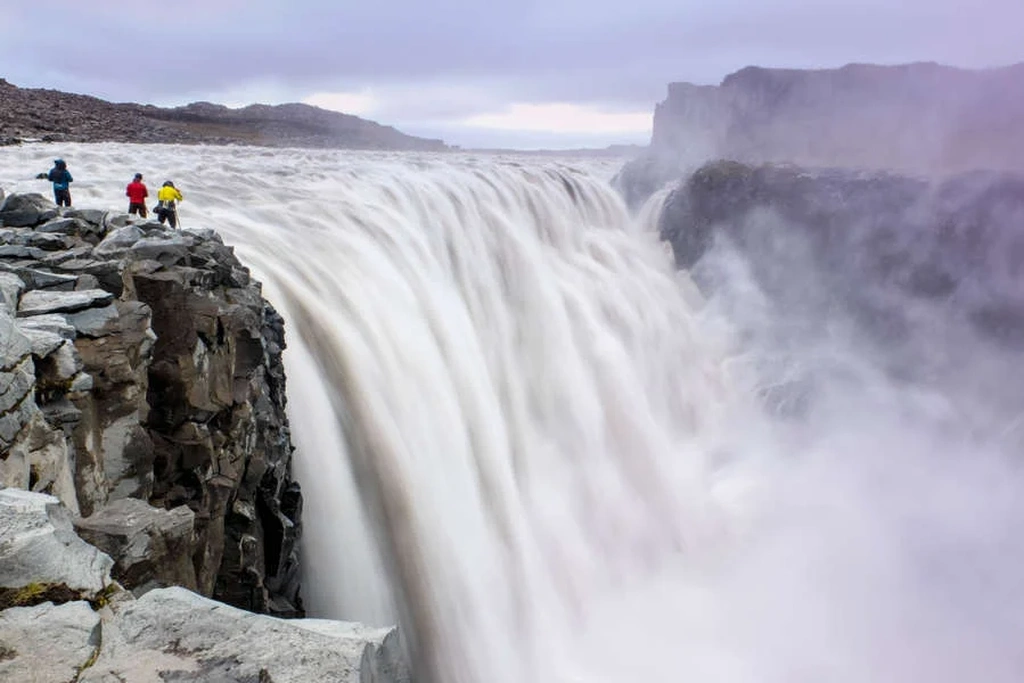 Tres personas con chaquetas coloridas están de pie al borde de un acantilado rocoso, observando la poderosa cascada Dettifoss en Islandia. La enorme cascada se precipita en el cañón nebuloso abajo, creando una escena dramática e impresionante. El cielo está nublado, añadiendo una atmósfera sombría al paisaje.