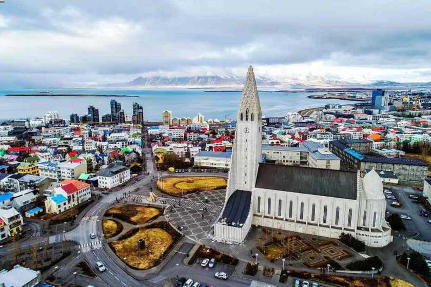 Vista aérea de Reykjavik, Islandia, con la icónica iglesia Hallgrímskirkja en primer plano, edificios coloridos y la costa con montañas al fondo bajo un cielo nublado.