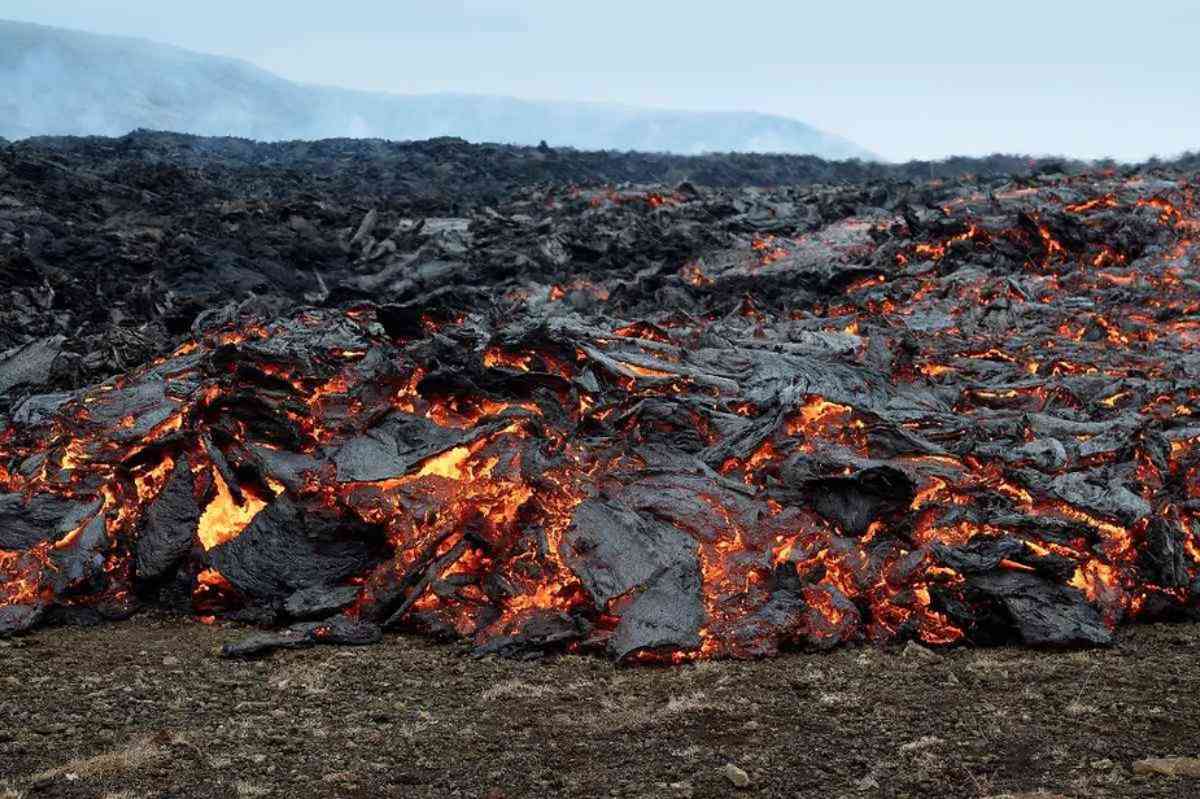 Primer plano de un flujo de lava activo en Islandia. La superficie está cubierta de roca de lava oscura y endurecida, con lava fundida de color naranja y rojo brillante visible entre las grietas. La lava parece avanzar lentamente y el calor emana de las áreas brillantes. El fondo muestra un paisaje de campos de lava enfriada que se extienden a lo lejos bajo un cielo nublado.