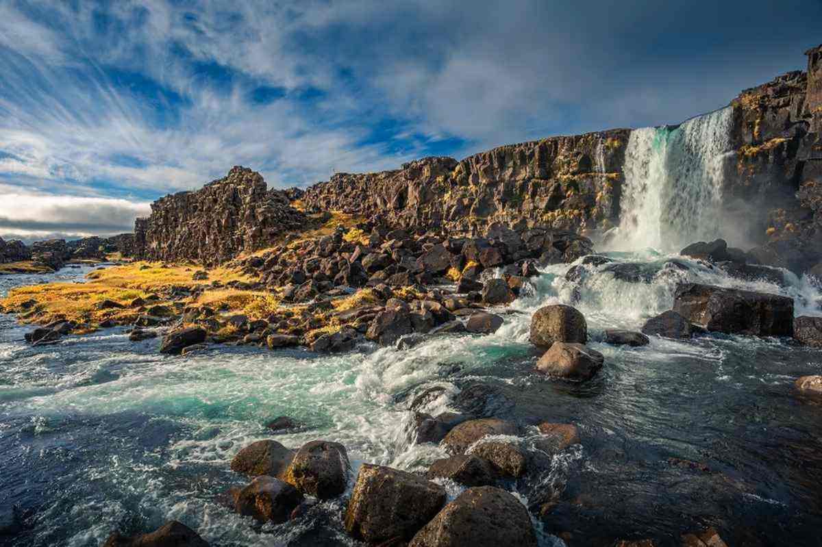 Vista panorámica de la cascada de Öxarárfoss cayendo sobre acantilados rocosos en el Parque Nacional de Thingvellir, Islandia, rodeada de terreno escarpado y vibrantes colores otoñales.