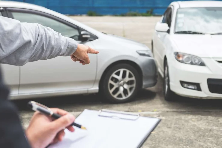 Una persona sosteniendo un portapapeles en primer plano mientras otra señala los daños entre un coche plateado y uno blanco tras una colisión menor.