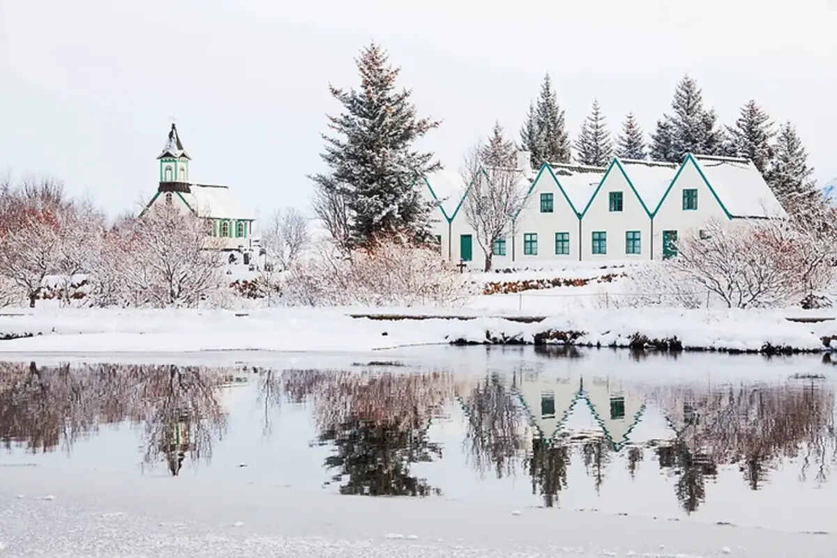 Maisons blanches et église traditionnelle islandaise recouvertes de neige, se reflétant dans un lac calme.