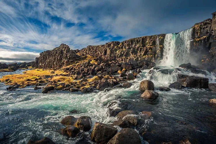 Vue panoramique de la cascade d'Öxarárfoss se jetant sur des falaises rocheuses dans le parc national de Thingvellir, en Islande, entourée d'un terrain accidenté et de couleurs automnales éclatantes.