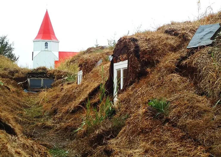  Maisons en tourbe typiques d'Islande entourées de végétation, avec une église au toit rouge en arrière-plan.