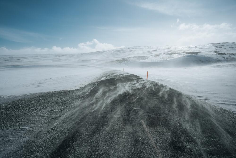 Une route isolée en Islande balayée par un vent glacial et de la neige tourbillonnante, entourée de collines enneigées sous un ciel clair.
