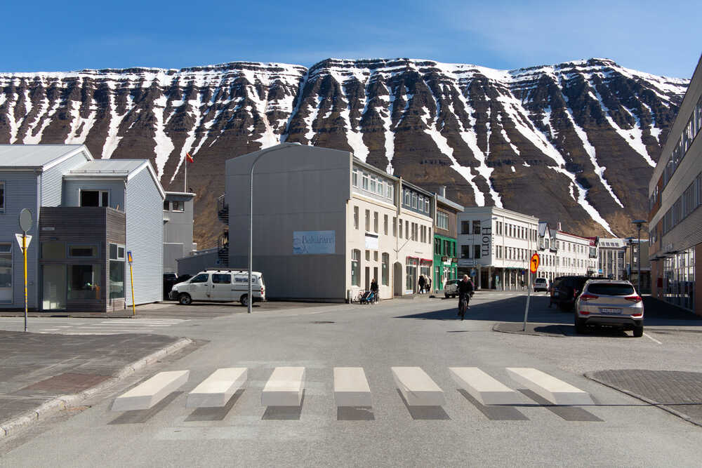 Passage piéton tridimensionnel dans la ville d'Ísafjörður, avec des montagnes enneigées en arrière-plan, offrant un paysage naturel spectaculaire.