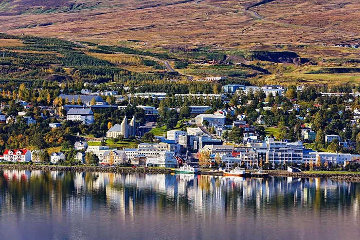 Vue panoramique de la ville d'Akureyri, avec des maisons colorées et des bâtiments bordant un fjord, entourée de collines verdoyantes et de montagnes au loin