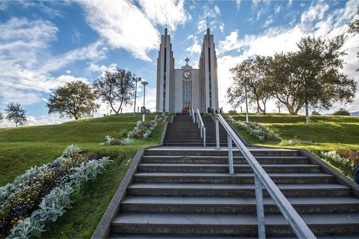 Église moderne en béton à Akureyri, avec des marches bordées de fleurs colorées menant à l'entrée principale sous un ciel bleu partiellement nuageux.