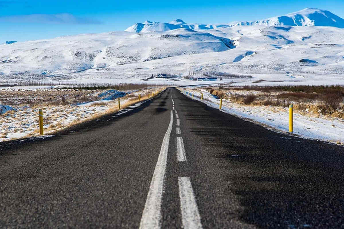 Une route droite entourée d'un paysage enneigé, avec des montagnes blanches au loin sous un ciel bleu éclatant.